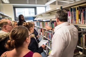 A group of people are gathered around a row of book shelving in a library. The figure on the right is giving instruction on finding items within the collection of items.