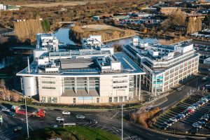 An aerial view of The Hub campus taken with the setting sun casting glowing light across the building front. Roads and roundabouts surround the buildings with traffic moving around.