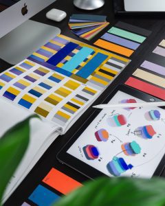 A range of colour swatches laid on a table. A book is open with swathes of yellow and blue options. Some colour sample pieces of card are laid next to it. In the lower corner, a digital tablet device and stylus pen are shown with more colour options drawn on the screen