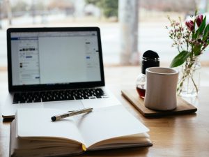 View from a table with a light window behind. A laptop sits at the rear of the table, with fresh coffee to the right. In the foreground is an open journal or notebook with a pen resting on the open pages.