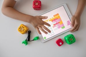 A child's hands interacting with a digital tablet device, playing a word game on the screen. The area surrounding the activity has random objects placed including large dice