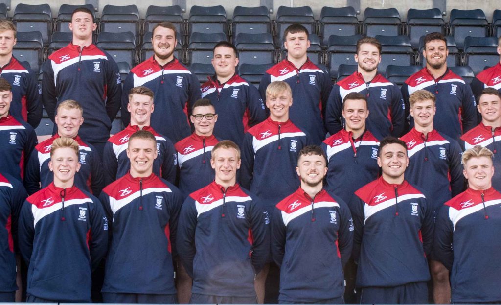 Photograph of the team lineup for the Rugby Academy, with all players smiling at the camera in group formation within a stadium seating area