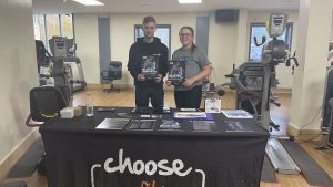 2 sports students standing behind a table in a gym holding promotional brochures