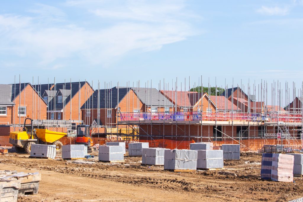 A construction site for a residential development is shown, with several brick houses in various stages of completion. The foreground features a muddy area with stacks of building materials and construction equipment, including a yellow dumper truck and an orange excavator. Scaffolding is erected around some of the houses, indicating ongoing construction. The scene is set under a bright blue sky.