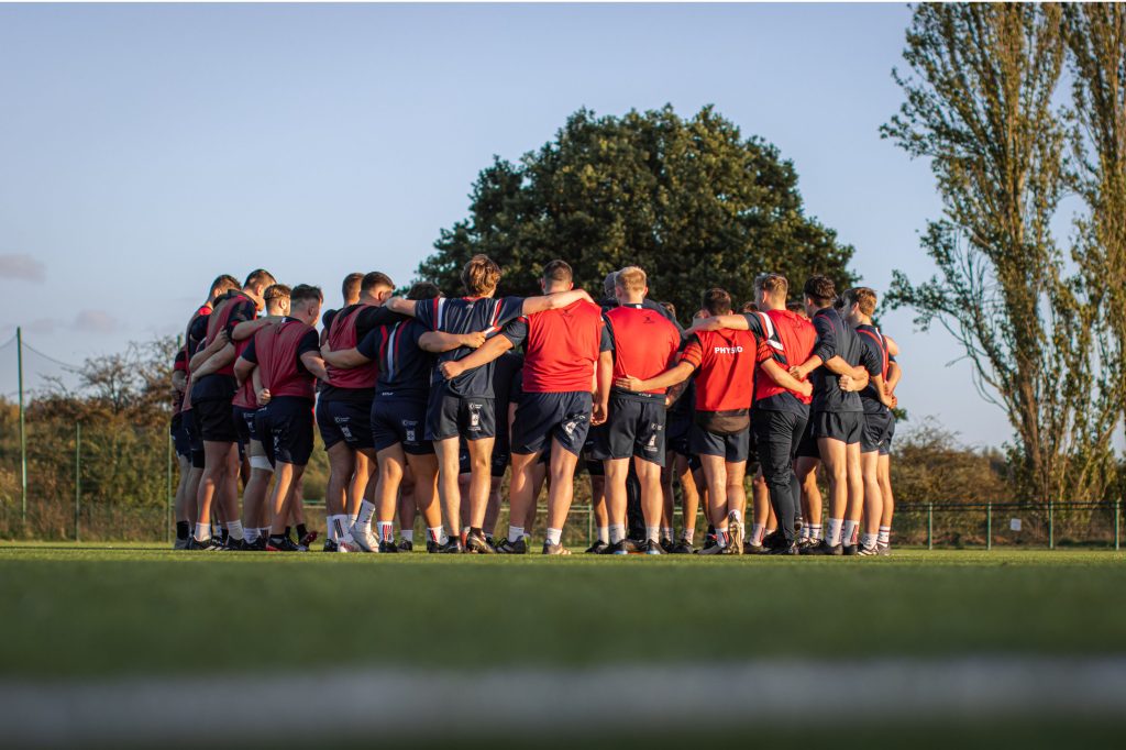 A group of athletes standing in a huddle on a grassy field. They are wearing sports attire, mostly consisting of navy blue shorts and red or blue jerseys. The players have their arms around each other's shoulders, indicating a team huddle or pep talk. In the background, there is a large tree, with two tall trees to the right. The setting appears to be during late afternoon or early evening, with clear skies and warm sunlight casting a golden glow on the scene. The players' backs are facing the camera, and they appear to be preparing for a game or practice session.