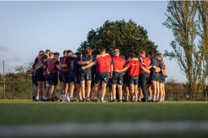 A group of athletes standing in a huddle on a grassy field. They are wearing sports attire, mostly consisting of navy blue shorts and red or blue jerseys. The players have their arms around each other's shoulders, indicating a team huddle or pep talk. In the background, there is a large tree, with two tall trees to the right. The setting appears to be during late afternoon or early evening, with clear skies and warm sunlight casting a golden glow on the scene. The players' backs are facing the camera, and they appear to be preparing for a game or practice session.