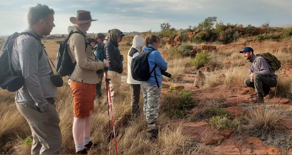 A group of people on a guided nature walk stand on a grassy, rocky terrain, listening to a guide who is crouched down explaining something. The participants are dressed in outdoor gear with backpacks, jackets, and hats, some holding cameras and walking sticks. The landscape is dry with tall grass, red rocks, and scattered shrubs under a partly cloudy sky.