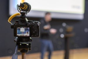 A close-up view of a professional camera mounted on a tripod with a yellow coiled microphone attached. The camera screen shows a live view of a presentation slide with charts and text. In the blurred background, a person is standing near a podium, gesturing while speaking. The setting is a lecture hall with a large projection screen.
