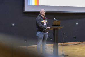 A speaker stands at a podium with a laptop and microphone, delivering a presentation in front of a large screen displaying colorful vertical stripes on the left side. The person is wearing a dark hoodie with a rainbow stripe design and jeans. The wooden floor and dark walls of the lecture hall are visible, with some foreground blur suggesting the image was taken from behind an audience member.