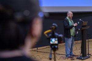 A male speaker stands at a podium equipped with a laptop and microphone, presenting in front of a large projection screen with a blue background. The person is wearing a colorful patterned shirt under a dark jacket and jeans. In the foreground, a camera with a yellow microphone attachment is partially visible, aimed at the speaker. Multiple cables are spread across the wooden floor.