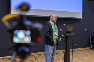 A males speaker stands at a podium equipped with a laptop and microphone, presenting in front of a large projection screen with a blue background. The person is wearing a colorful patterned shirt under a dark jacket and jeans. In the foreground, a camera with a yellow microphone attachment is partially visible, aimed at the speaker. Multiple cables are spread across the wooden floor.