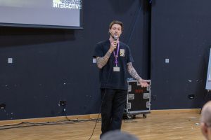 A male is speaking into a microphone while standing in front of a large screen in a lecture hall. The individual is wearing a black T-shirt with visible tattoos on both arms and a lanyard with an ID badge. Behind them, technical equipment cases and cables are placed against the wall. The wooden floor and dark walls of the hall are visible, along with part of the projection screen displaying text and graphics.