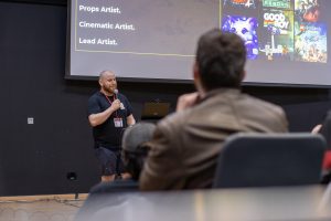 A male speaker is holding a microphone and presenting in front of a large projection screen displaying text and colorful game artwork. The visible text includes roles such as “Props Artist,” “Cinematic Artist,” and “Lead Artist.” Several game covers are shown on the right side of the screen, including vibrant illustrations. The speaker is wearing a black T-shirt and shorts with a lanyard and badge. Audience members are seated in the foreground, facing the speaker, with the lecture hall’s tiered seating partially visible.