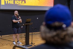 A person is presenting at a podium with a laptop and microphone in a lecture hall. The large screen behind them displays bold text reading “THE IDEA” on an orange textured background. The speaker is wearing a black T-shirt and shorts with a lanyard and badge. In the foreground, an audience member wearing a bright blue cap is visible, slightly out of focus.