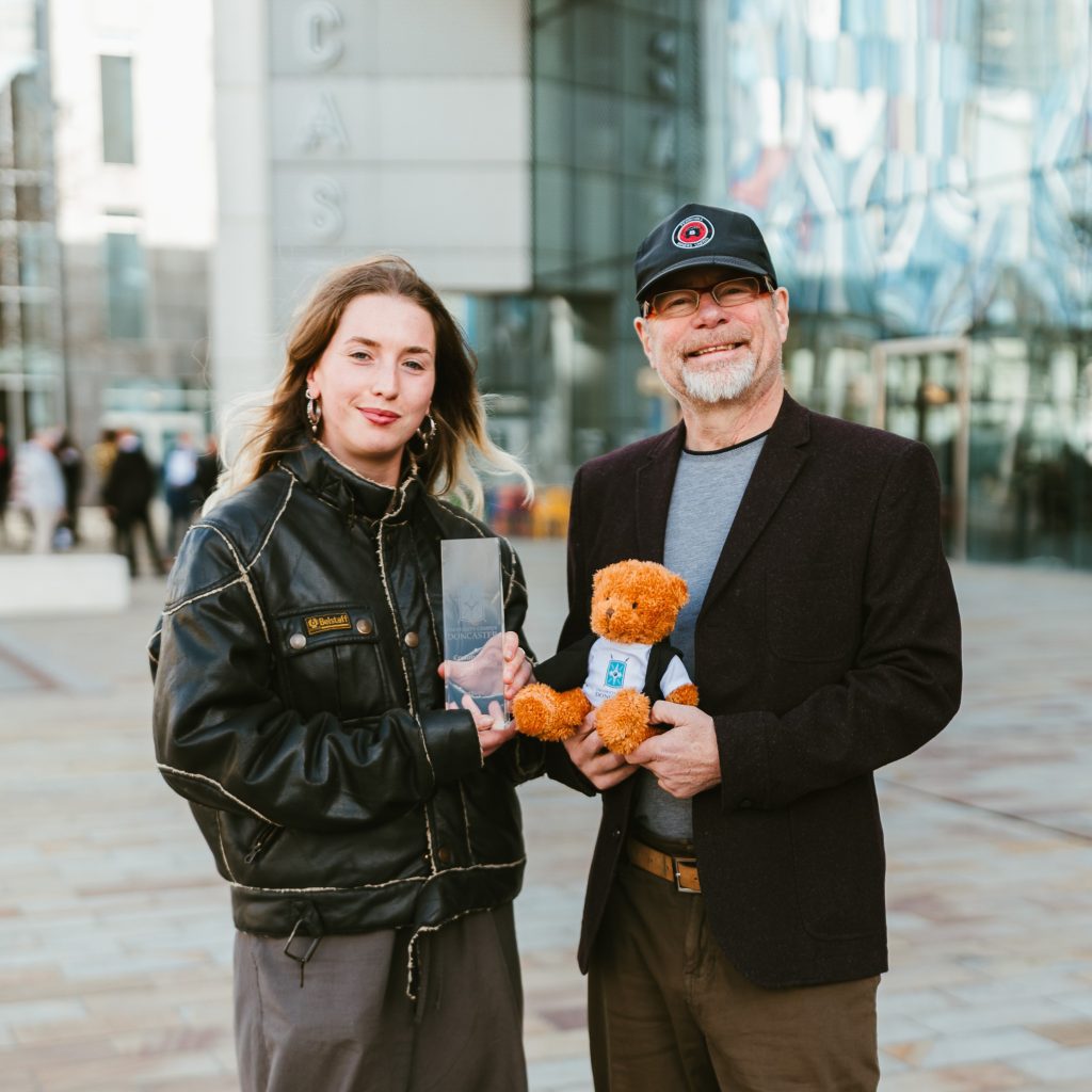 Two people stand together on a spacious, tiled plaza in front of a large modern building with tall glass panels and reflective surfaces. The person on the left is a young adult with light skin and long ombré hair, wearing a black leather jacket with prominent stitching, loose gray pants, and tan shoes. They hold a rectangular glass award with both hands and face the camera with a calm expression. The male on the right is an older adult with light skin, a gray beard, glasses, and a black baseball cap. They are dressed in a dark blazer over a gray shirt, brown pants, and dark shoes, smiling warmly while holding a small teddy bear in a white shirt with a blue emblem. People can be seen walking in the background near the building’s entrance, and sunlight reflects softly off the surrounding glass.