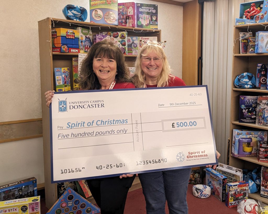 Two women stand side by side in a brightly lit room lined with shelves full of children’s toys. They are both smiling warmly as they hold a large ceremonial cheque between them. The woman on the left has medium-length dark hair with bangs, is wearing a black top, and has a red lanyard around her neck. The woman on the right has long, light-blonde hair and glasses, and is wearing a red sweater with a lanyard. The oversized cheque reads “University Campus Doncaster” at the top and is made payable to “Spirit of Christmas” for £500.00, dated 9th December 2025. Around them, neatly arranged toys fill the shelving—items like board games, dolls, art kits, action figures, sports sets, and boxed science toys. Some toys are stacked on the floor in front of the shelves. Small decorative string lights are woven along the upper shelving, adding a festive touch. The overall scene conveys generosity, community spirit, and preparation for holiday gift-giving.