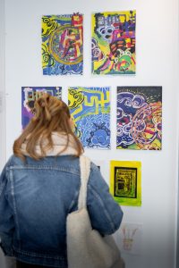 A woman with light brown hair wearing a denim jacket stands in front of a wall displaying nine colourful abstract paintings featuring swirls, geometric shapes, and layered patterns.
