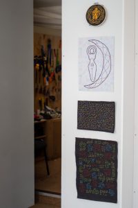 A vertical row of textile artworks on a white wall: a circular embroidered piece at the top, a stitched figure inside a crescent moon, and two geometric pattern textiles below. A workshop space is visible in the background.