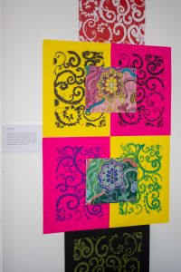 A vertical arrangement of textile artworks and prints featuring bold pink, yellow, green, and black backgrounds with ornate swirling patterns and a central floral design.