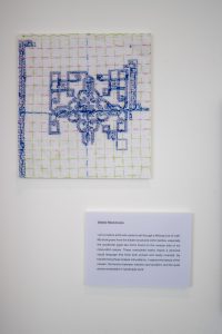 A stitched textile artwork on white fabric featuring a symmetrical blue pattern made with cross‑stitch. A printed artist statement is mounted on the wall below it.