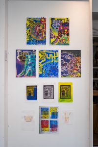 A gallery wall featuring nine colourful abstract prints arranged in a grid. The artworks include layered shapes, lines, and bright colours such as blue, yellow, and pink.