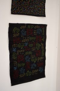 Two textile pieces in dark fabric, each stitched with bright, colourful geometric and maze‑like line patterns in red, green, blue, and yellow.