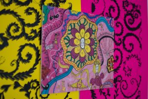 A close‑up of a pink textile artwork featuring an intricate floral and decorative design, mounted on a bold yellow and pink patterned background.
