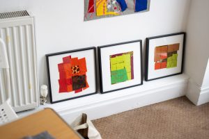 Three framed fabric artworks resting on the floor against a wall. Each features patchwork‑style compositions in warm colours such as orange, red, yellow, and green.