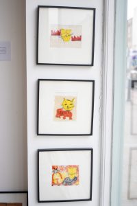 A vertical set of three framed prints showing stylised yellow cat illustrations. Each print uses different background colours and textures.