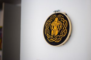 A circular embroidered artwork in a wooden hoop, showing a stylised human‑like figure outlined in yellow thread against a black background with swirling decorative borders.
