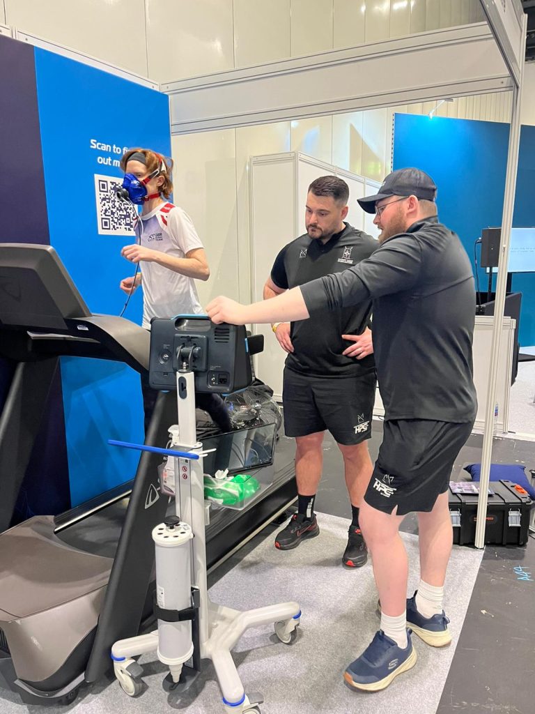 A person runs on a treadmill while wearing a blue-and-red breathing mask connected to testing equipment. Two staff members in black sportswear stand beside the treadmill, monitoring the performance device and adjusting its settings. The scene takes place in an indoor exhibition or testing area with blue display panels and equipment around them.