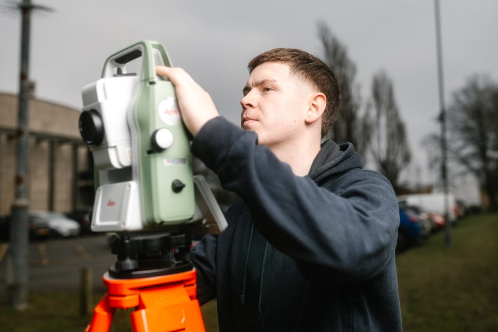 A you white male outdoors operates a large green and white Leica surveying instrument mounted on a bright orange tripod. The man is wearing a dark navy hoodie and is adjusting the device with both hands. The background includes blurred bare trees, parked cars, and a grey building under an overcast sky.
