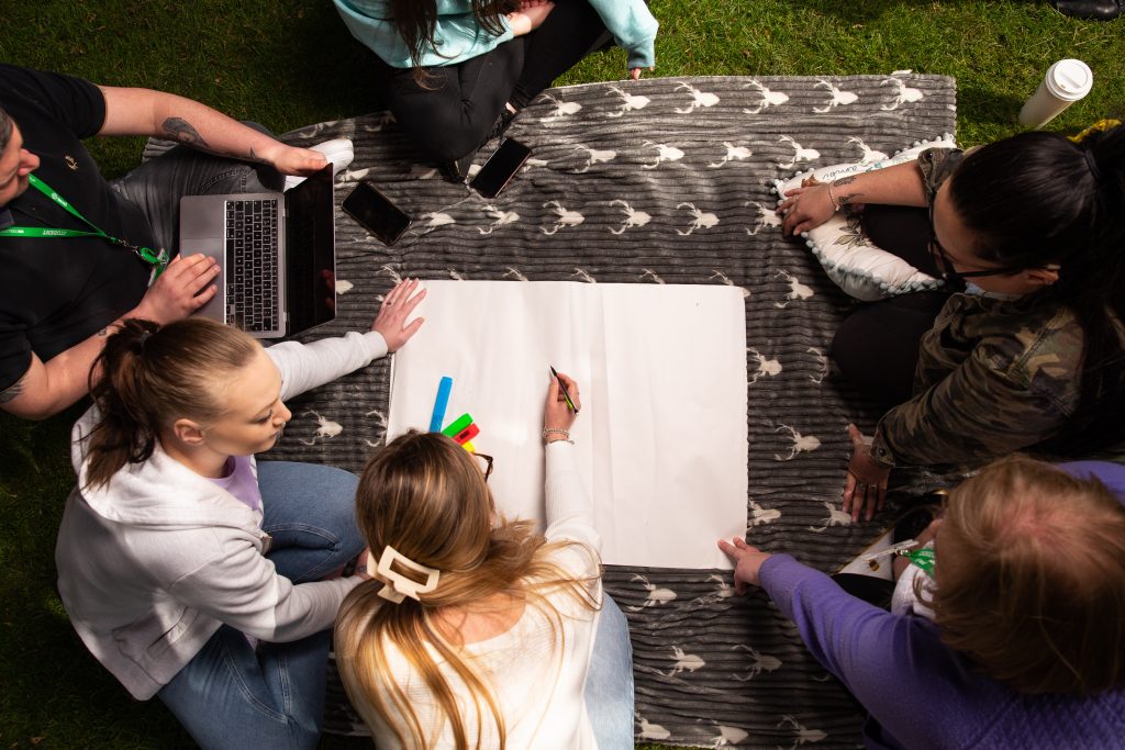 Group of students sitting on a blanket outdoors, collaborating around a large sheet of paper with markers, a laptop, and phones.