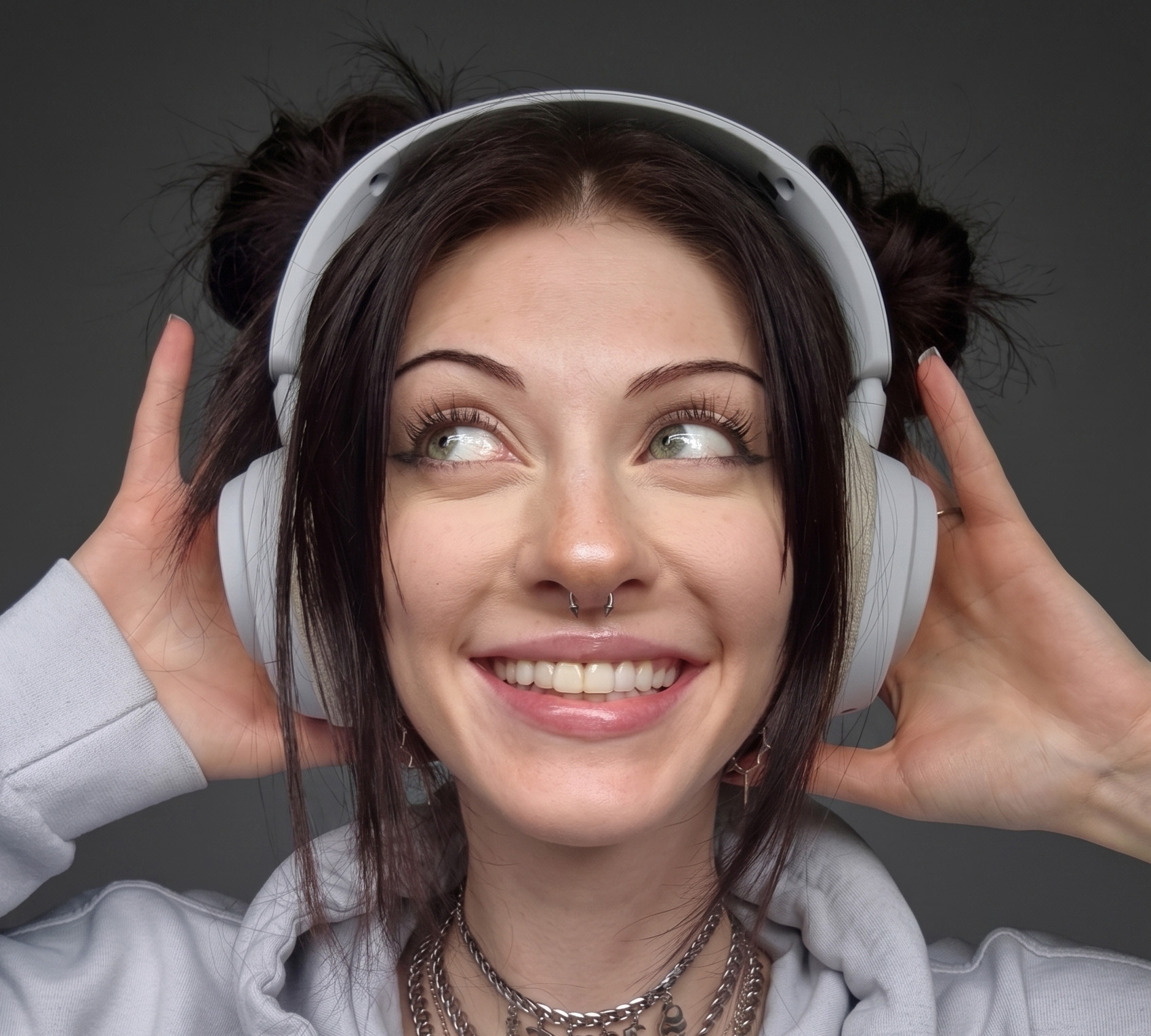 Close-up portrait of a person wearing large light-gray over-ear headphones. The person is centered in the frame and holds both sides of the headphones with their hands. Dark hair is styled with two small buns positioned behind the headphones, with longer strands framing the face. The person is wearing a light-gray hoodie and several layered silver chain necklaces. A septum piercing is visible, along with small dangling earrings. The person's eyes are looking upward and to the side rather than directly at the camera. The image is tightly cropped around the head and shoulders, filling most of the frame. The background is a plain, softly blurred dark gray surface with no visible objects or text. Soft, even lighting illuminates the face and headphones, creating a clean, professional appearance with clear detail in the hair, skin, jewelry, and fabric textures. The overall color palette is neutral, consisting mainly of gray, silver, black, and muted tones.