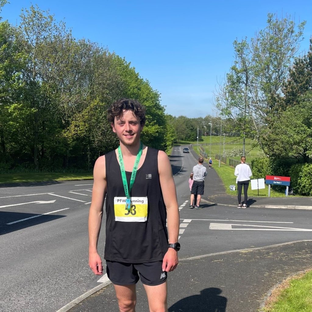 Young male athlete wearing a black vest sports vest and shorts is standing beside a road with a green and gold winners medal around his neck.