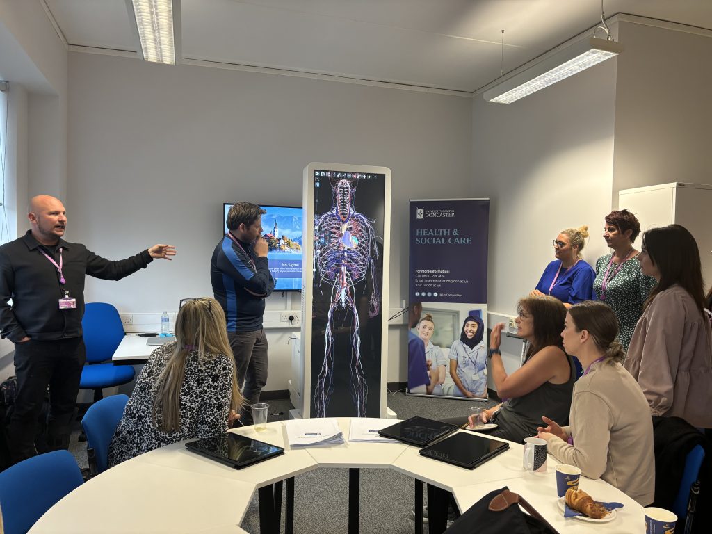 The image shows a group of people in a classroom setting. They are gathered around a digital display that shows an anatomical diagram of the human body. One person is standing and gesturing while speaking, possibly explaining something to the group. Another person stands near the display, looking at it thoughtfully. Others are seated at a table with tablets, cups of coffee, and pastries. A banner in the background reads "HEALTH & SOCIAL CARE" and mentions "University Campus Doncaster." The atmosphere suggests a professional development or educational session related to health and social care.