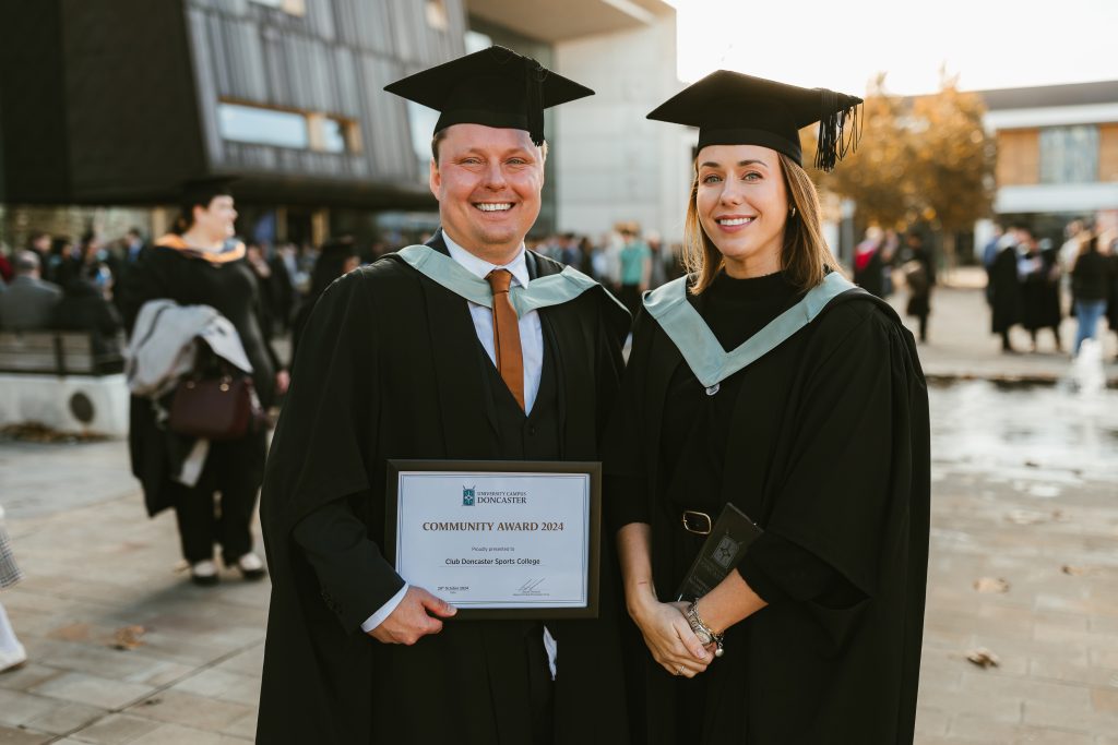 The two individuals in the foreground are dressed in traditional academic dress, including black gowns with light green stoles and graduation caps (mortarboards). The individual on the left: A smiling man, holding a framed award titled "Community Award 2024," presented to "Club Doncaster Sports College." He is wearing a suit and tie under the graduation attire, and his cheerful expression reflects pride and accomplishment. The individual on the right: A smiling woman, also in graduation regalia. She is holding what appears to be a trophy or plaque in her hand. Her outfit under the gown includes a black top, and she accessorizes with a belt and bracelets. Her posture and smile convey happiness and confidence. The background features a modern building, with a crowd of other graduates, family members, and attendees engaging in celebratory activities. The light suggests it’s daytime, with autumn leaves scattered on the ground, hinting at a fall graduation season.