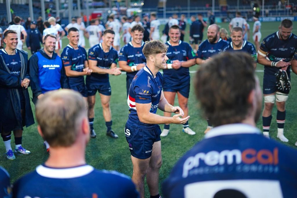 A group of rugby players in dark blue uniforms stand in a circle on a pitch, clapping and smiling. The central focus is a young male player with short brown hair, wearing a blue rugby kit with logos and white-red detailing on the sleeves. He is standing in the middle of the circle, slightly bent forward, with his hands together as if addressing or being celebrated by his teammates. The players surrounding him are laughing, smiling, and clapping, creating a joyful and supportive atmosphere. In the background, more players in white kits are seen walking away, and stadium seating with spectators is visible. The scene appears to be post-match, with a sense of camaraderie and celebration among the team.