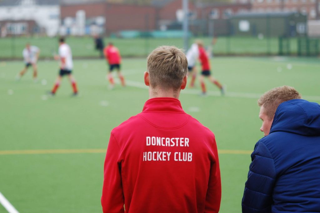 Two men watching a field hockey match; one is wearing a red jacket with 'Doncaster Hockey Club' written on the back, while players in red and white uniforms are seen blurred in action on the green turf field."