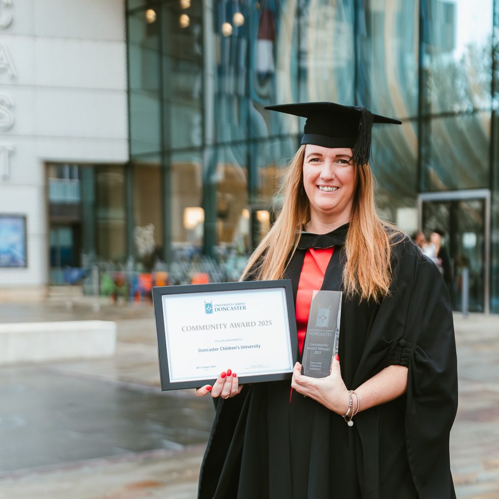 A woman with long brown hair in a black gown and mortarboard with a red stole stands outdoors in front of a modern glass building. The person holds a framed certificate titled “Community Award 2025” from Doncaster Children’s University in the left hand and a clear rectangular trophy in the right hand. The background includes large reflective glass panels, an entrance door, and some outdoor seating with plants. The ground appears wet, suggesting recent rain.