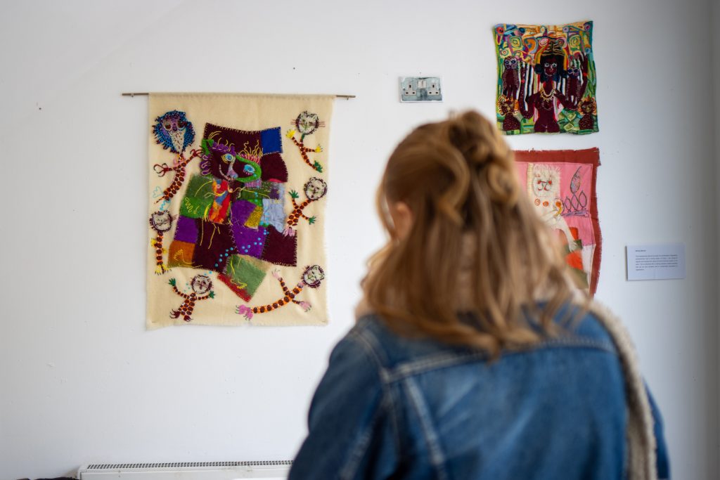 A person with light brown hair wearing a denim jacket stands viewing three textile artworks on a white wall, including a large stitched piece with vivid colours and abstract shapes.